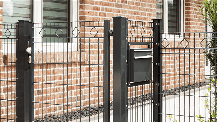 Et nærbillede af et sort metaltrådshegn med Panelhegn "MORITS" 2D 6/5/6 mm med diamantformede mønstre og en matchende port foran et hus med murstensvægge og hvidindrammede vinduer. Porten har en lås og en sort postkasse monteret på.