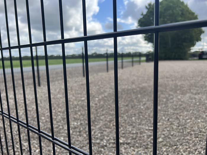 Nærbillede af Panelhegn 2D 6/5/6 mm Pulverlakeret Sort - Komplet Inkl. Stolper & Beslag, et slankt, sort metal hegnspanel, mod en grusoverflade med et træ i baggrunden under en delvist overskyet himmel. Perspektivet skaber en følelse af dybde og fremhæver hegnspaneler og træer, der strækker sig i det fjerne.