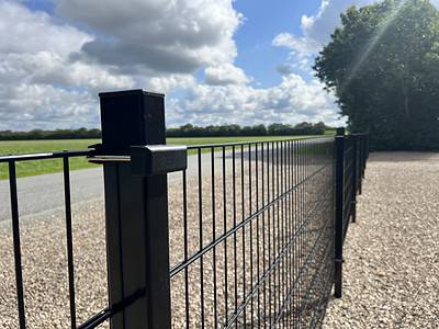 Et nærbillede af et Panelhegn 2D 6/5/6 mm Pulverlakeret Sort hegn, med lodrette og vandrette stænger, sat på en grusbelagt grund. Panelet er placeret i højre side og strækker sig mod et lyst, solrigt landskab med grønne marker, en vej og en blå himmel fyldt med hvide skyer. Dette komplette sæt inkluderer stolper og beslag for nem installation.