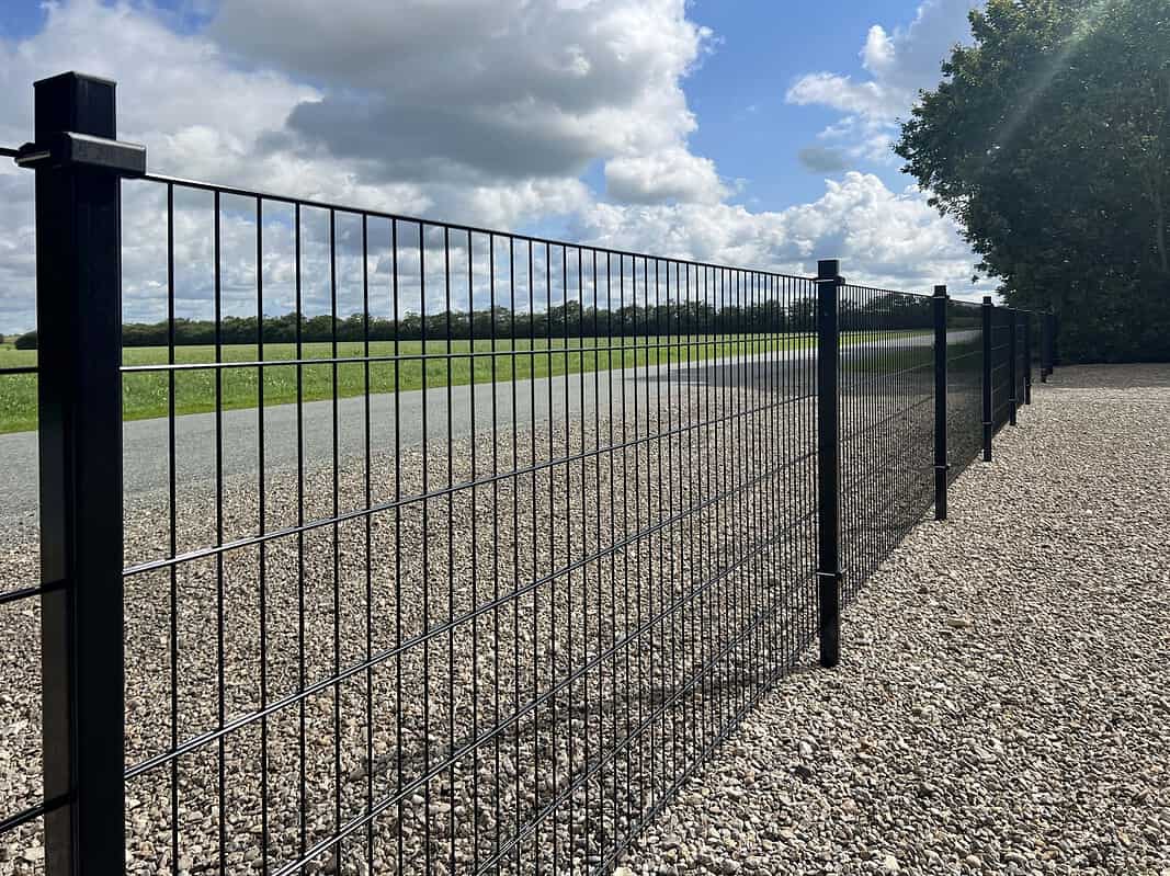 En lang Panelhegn 2D 6/5/6 mm Pulverlakeret Sort - Komplet Inkl. Stolper & Beslag hegn strækker sig langs en asfalteret vej. Hegnet står på en grusoverflade, med græs og træer synlige i baggrunden under en delvist overskyet himmel.