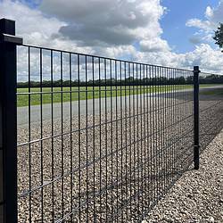 En lang Panelhegn 2D 6/5/6 mm Pulverlakeret Sort - Komplet Inkl. Stolper & Beslag hegn strækker sig langs en asfalteret vej. Hegnet står på en grusoverflade, med græs og træer synlige i baggrunden under en delvist overskyet himmel.