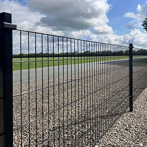 En lang Panelhegn 2D 6/5/6 mm Pulverlakeret Sort - Komplet Inkl. Stolper & Beslag hegn strækker sig langs en asfalteret vej. Hegnet står på en grusoverflade, med græs og træer synlige i baggrunden under en delvist overskyet himmel.