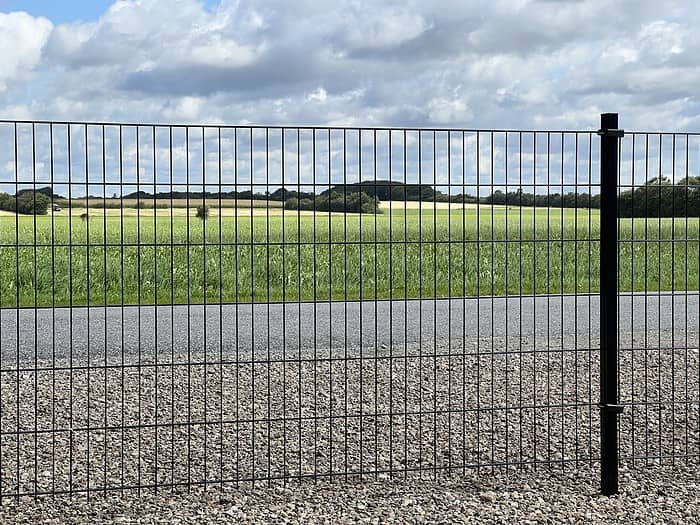 Et sort metalhegn – konstrueret med Panelhegn 2D 6/5/6 mm Pulverlakeret Sort, komplet med stolper og beslag – strækker sig vandret hen over grusstien i forgrunden. Ud over Panelhegn er der asfalteret vej, grønne marker, fjerne træer og en delvis skyet himmel.