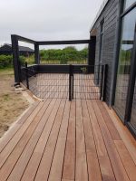 En moderne udendørs terrasse med træterrasse, omkranset af sort trådhegn og en lækker Havelåge 2D Panelfyld 8/6/8mm Rammeløs "BLACKY" Komplet port. Terrassen er knyttet til en mørk bygning med store glasdøre. Det omkringliggende område omfatter græs og buske under en overskyet himmel.