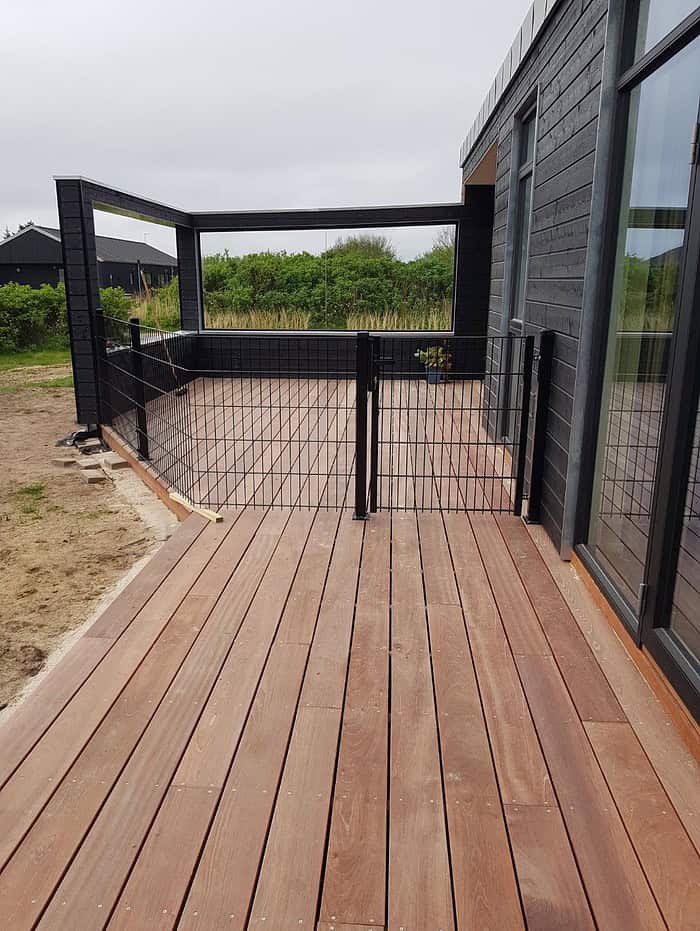 En moderne udendørs terrasse med træterrasse, omkranset af sort trådhegn og en lækker Havelåge 2D Panelfyld 8/6/8mm Rammeløs "BLACKY" Komplet port. Terrassen er knyttet til en mørk bygning med store glasdøre. Det omkringliggende område omfatter græs og buske under en overskyet himmel.