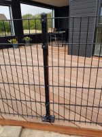 Et sort metalhegn med lodrette og vandrette stænger, identificeret som "Havelåge 2D Panelfyld 8/6/8mm Rammeløs 'BLACKY' Komplet," er monteret på et trædæk, der er en del af et moderne, mørkfarvet hus. Glasskydedøre fører til rammeløs låge, med nogle potteplanter synlige i baggrunden.