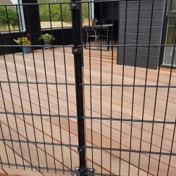 Et sort metalhegn med lodrette og vandrette stænger, identificeret som "Havelåge 2D Panelfyld 8/6/8mm Rammeløs 'BLACKY' Komplet," er monteret på et trædæk, der er en del af et moderne, mørkfarvet hus. Glasskydedøre fører til rammeløs låge, med nogle potteplanter synlige i baggrunden.