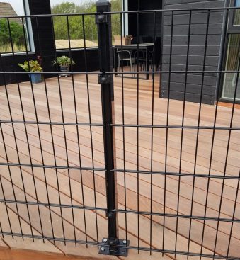 Et sort metalhegn med lodrette og vandrette stænger, identificeret som "Havelåge 2D Panelfyld 8/6/8mm Rammeløs 'BLACKY' Komplet," er monteret på et trædæk, der er en del af et moderne, mørkfarvet hus. Glasskydedøre fører til rammeløs låge, med nogle potteplanter synlige i baggrunden.