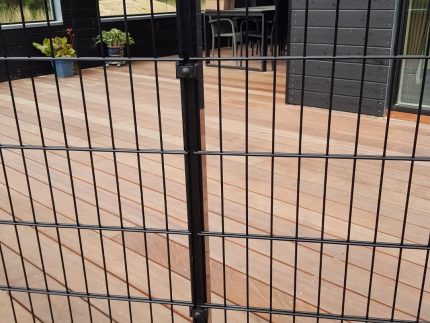 Et sort metalhegn med lodrette og vandrette stænger, identificeret som "Havelåge 2D Panelfyld 8/6/8mm Rammeløs 'BLACKY' Komplet," er monteret på et trædæk, der er en del af et moderne, mørkfarvet hus. Glasskydedøre fører til rammeløs låge, med nogle potteplanter synlige i baggrunden.