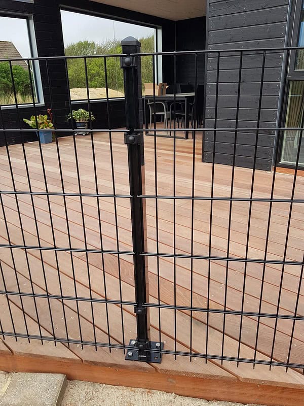 Et sort metalhegn med lodrette og vandrette stænger, identificeret som "Havelåge 2D Panelfyld 8/6/8mm Rammeløs 'BLACKY' Komplet," er monteret på et trædæk, der er en del af et moderne, mørkfarvet hus. Glasskydedøre fører til rammeløs låge, med nogle potteplanter synlige i baggrunden.