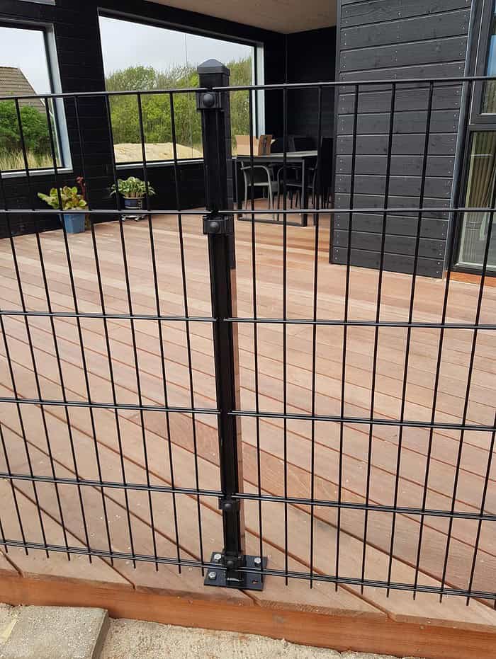 Et sort metalhegn med lodrette og vandrette stænger, identificeret som "Havelåge 2D Panelfyld 8/6/8mm Rammeløs 'BLACKY' Komplet," er monteret på et trædæk, der er en del af et moderne, mørkfarvet hus. Glasskydedøre fører til rammeløs låge, med nogle potteplanter synlige i baggrunden.