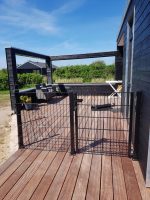 Et solrigt træterrasse knyttet til et sort hus med udendørsmøbler inklusive en polstret sofa og en hvid foldbar stol. Dækket omsluttes af den slanke Havelåge 2D Panelfyld 8/6/8mm Rammeløs "BLACKY" Komplet port. I baggrunden er der grønne buske og yderligere strukturer under en klar blå himmel.