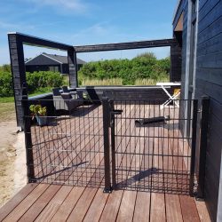 Et solrigt træterrasse knyttet til et sort hus med udendørsmøbler inklusive en polstret sofa og en hvid foldbar stol. Dækket omsluttes af den slanke Havelåge 2D Panelfyld 8/6/8mm Rammeløs "BLACKY" Komplet port. I baggrunden er der grønne buske og yderligere strukturer under en klar blå himmel.