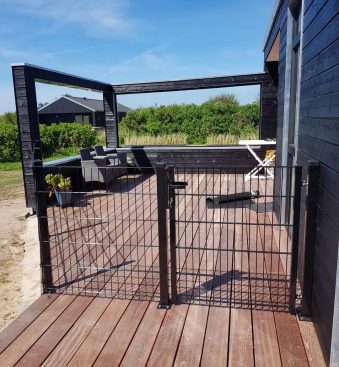 Et solrigt træterrasse knyttet til et sort hus med udendørsmøbler inklusive en polstret sofa og en hvid foldbar stol. Dækket omsluttes af den slanke Havelåge 2D Panelfyld 8/6/8mm Rammeløs "BLACKY" Komplet port. I baggrunden er der grønne buske og yderligere strukturer under en klar blå himmel.