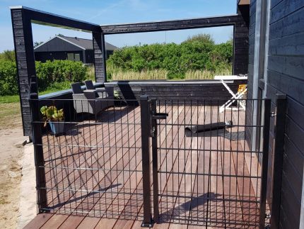 Et solrigt træterrasse knyttet til et sort hus med udendørsmøbler inklusive en polstret sofa og en hvid foldbar stol. Dækket omsluttes af den slanke Havelåge 2D Panelfyld 8/6/8mm Rammeløs "BLACKY" Komplet port. I baggrunden er der grønne buske og yderligere strukturer under en klar blå himmel.