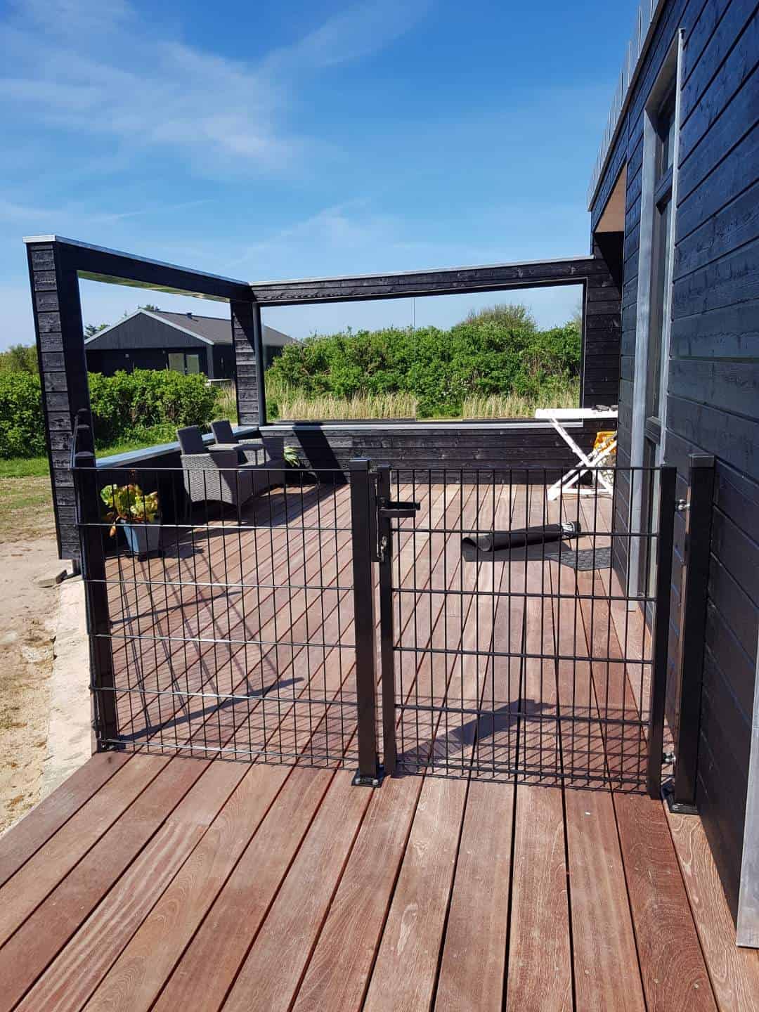Et solrigt træterrasse knyttet til et sort hus med udendørsmøbler inklusive en polstret sofa og en hvid foldbar stol. Dækket omsluttes af den slanke Havelåge 2D Panelfyld 8/6/8mm Rammeløs "BLACKY" Komplet port. I baggrunden er der grønne buske og yderligere strukturer under en klar blå himmel.