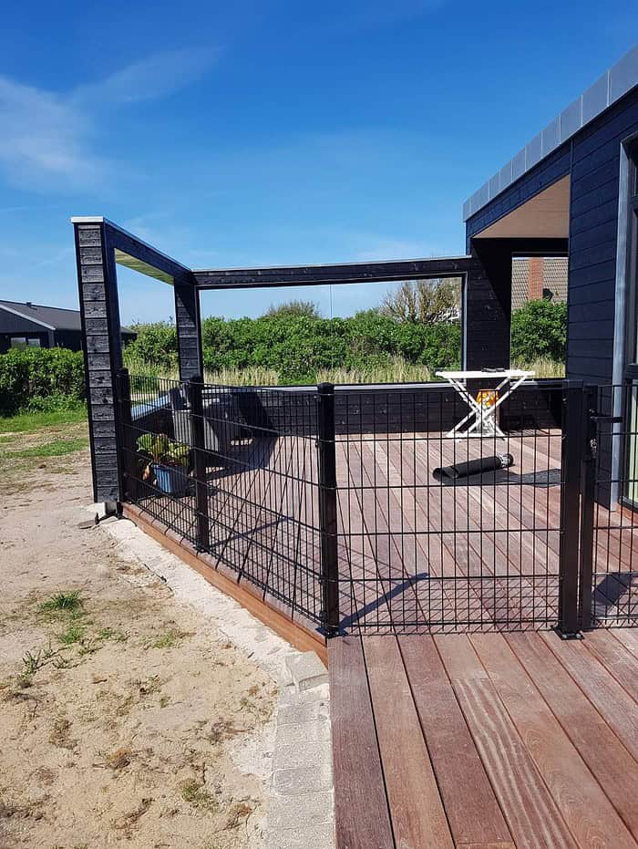 Et indhegnet træterrasse knyttet til et mørkfarvet hus har en pergola med åben ramme. Indenfor det indhegnede område er der en Havelåge 2D Panelfyld 8/6/8mm Rammeløs "BLACKY" Komplet, en potteplante og et hvidt udklapsbord med gult mønster. Baggrunden inkluderer en græsklædt gård og klar blå himmel.