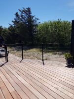 Et solrigt udendørs dæk med trægulv og et sort metalhegn, der omslutter området. Ud over hegnet er der en sandet gård med spredte græspletter, omkranset af høje grønne træer under en klar blå himmel. En slank Havelåge 2D Panelfyld 8/6/8mm Rammeløs "BLACKY" Komplet tilføjer elegance til scenen, mens en potteplante er placeret i hjørnet af dækket.