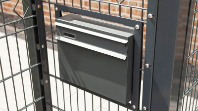 En lækker sort postkasse med et moderne design er monteret på et Panelhegn "MORITS" 2D 6/5/6 mm metalhegn. Hegnets geometriske mønster skiller sig ud med en baggrund, der inkluderer en murstensvæg og lyse belægningssten. Postkassen har en bred spalte og et lille håndtag for nem adgang.