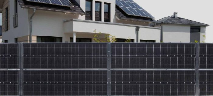 Moderne hus udstyret med solcellepaneler på taget, omgivet af en høj, mørkfarvet metalhegn. Bygningen har et rent, moderne design med store vinduer og fladt tag. Billedet fremhæver vedvarende energi og moderne boligarkitektur.