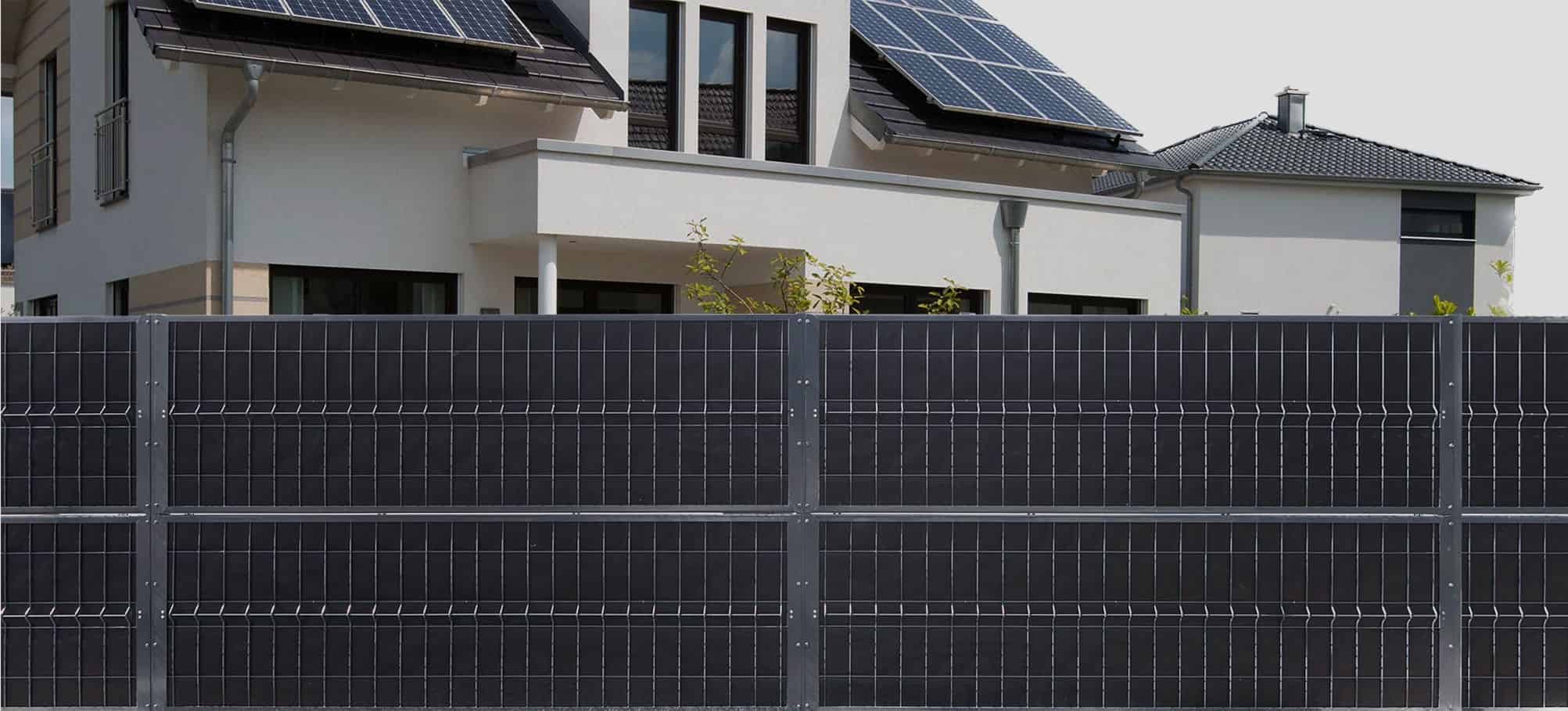 Moderne hus udstyret med solcellepaneler på taget, omgivet af en høj, mørkfarvet metalhegn. Bygningen har et rent, moderne design med store vinduer og fladt tag. Billedet fremhæver vedvarende energi og moderne boligarkitektur.