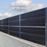 Et højt sort metalhegn med lodrette og vandrette stænger står langs en asfalteret vej. Himlen er delvist overskyet, og lidt grus kanter bunden af hegnet. Denne Støj Hegn "ECO" 18Db - 3D Panel Pulverlakeret Metal - Komplet Inkl. stolper fremstår robust og ensartet fordelt, hvilket sikrer både privatliv og støjreduktion for husejere.