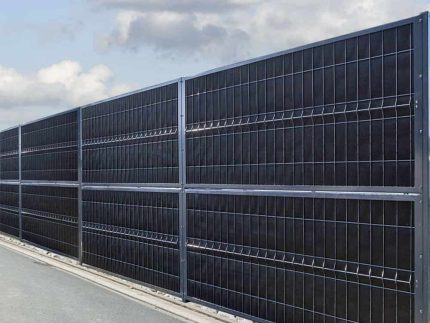 Et højt sort metalhegn med lodrette og vandrette stænger står langs en asfalteret vej. Himlen er delvist overskyet, og lidt grus kanter bunden af hegnet. Denne Støj Hegn "ECO" 18Db - 3D Panel Pulverlakeret Metal - Komplet Inkl. stolper fremstår robust og ensartet fordelt, hvilket sikrer både privatliv og støjreduktion for husejere.