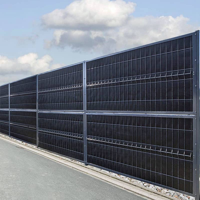 Et højt sort metalhegn med lodrette og vandrette stænger står langs en asfalteret vej. Himlen er delvist overskyet, og lidt grus kanter bunden af hegnet. Denne Støj Hegn "ECO" 18Db - 3D Panel Pulverlakeret Metal - Komplet Inkl. stolper fremstår robust og ensartet fordelt, hvilket sikrer både privatliv og støjreduktion for husejere.