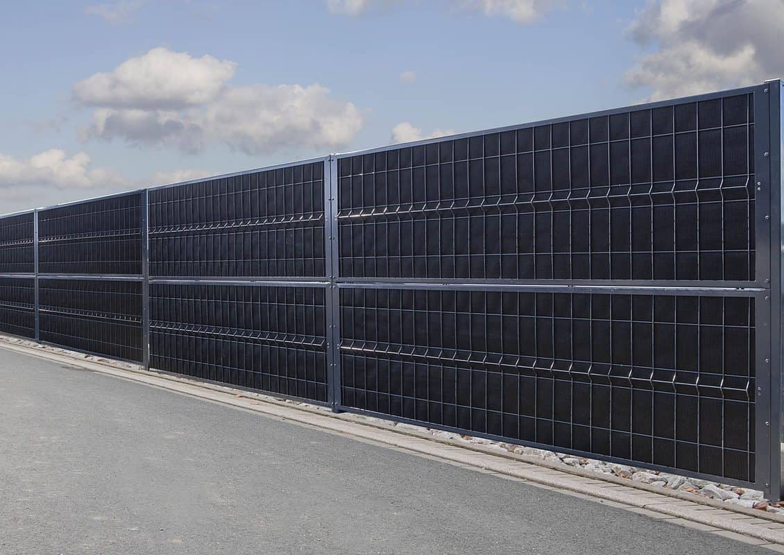 Et langt sort solpanelhegn, der ligner en støjhegn, er monteret langs en grå asfalteret vej med en overskyet himmel i baggrunden. Panelerne er opdelt i sektioner og monteret på en metalramme med sten i bunden, hvilket skaber en imponerende forside til vejkanten.