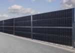 Et langt sort solpanelhegn, der ligner en støjhegn, er monteret langs en grå asfalteret vej med en overskyet himmel i baggrunden. Panelerne er opdelt i sektioner og monteret på en metalramme med sten i bunden, hvilket skaber en imponerende forside til vejkanten.