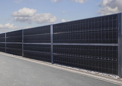 Et langt sort støjhegn, er monteret langs en grå asfalteret vej med en overskyet himmel i baggrunden. Panelerne er opdelt i sektioner og monteret på en metalramme med sten i bunden, hvilket skaber en imponerende forside til vejkanten.