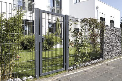 Omslutter den velholdte have ligger Panelhegn 2D 6/5/6 mm Pulverlakeret Sort - Komplet Inkl. Stolper & Beslag, som har en kombination af metal- og stensektioner. Bag denne sofistikerede panelstruktur ligger der en plet af frodigt grønt græs, buske og en hvid bygning med store vinduer og altaner.