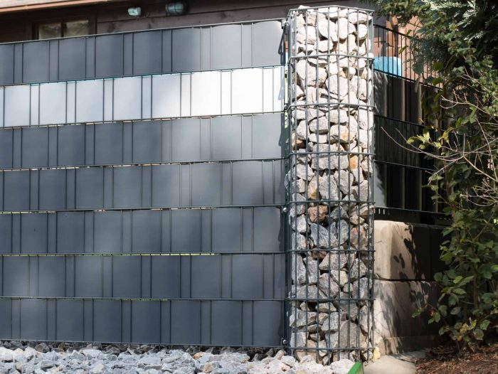 En moderne bygningsfacade har en kombination af flad, grå Gabionhegn – Panelhegn “ONYX” 2D OVERFL.Behandl? paneler og et cylindrisk trådbur fyldt med sten. Jorden er dækket af ensartede, lysegrå sten, og en grøn busk er synlig i højre side af billedet.