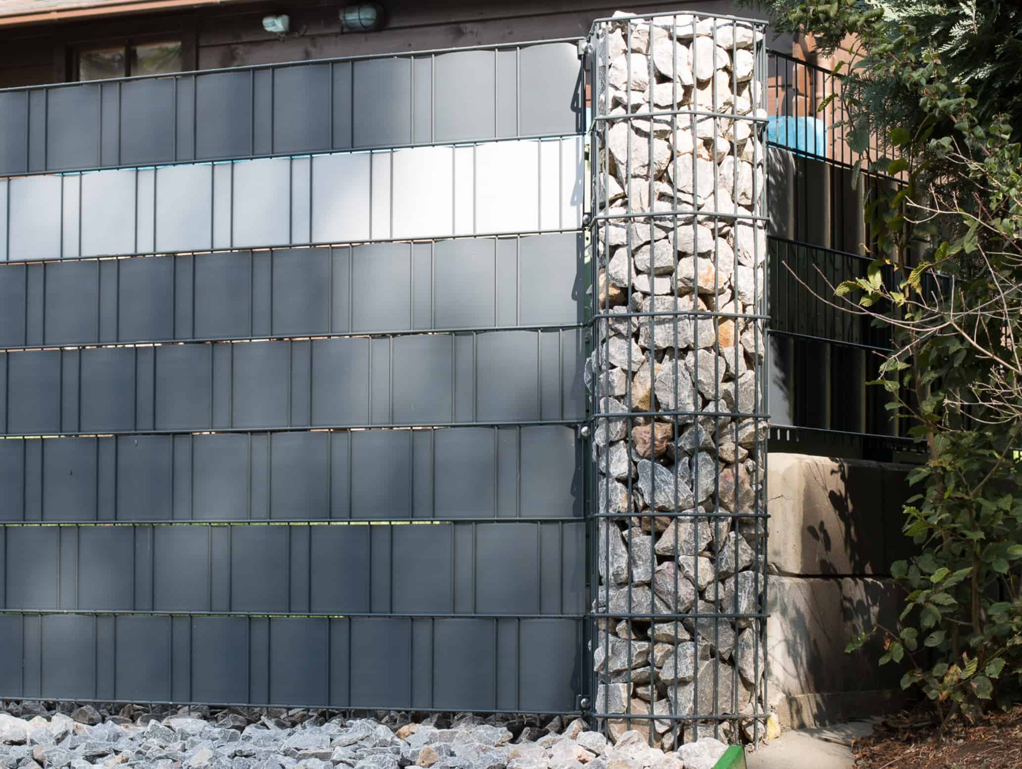 En moderne bygningsfacade har en kombination af flad, grå Gabionhegn – Panelhegn “ONYX” 2D OVERFL.Behandl? paneler og et cylindrisk trådbur fyldt med sten. Jorden er dækket af ensartede, lysegrå sten, og en grøn busk er synlig i højre side af billedet.