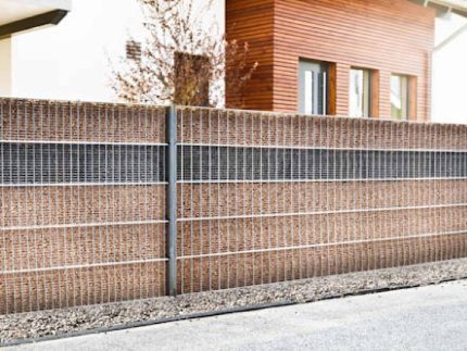 En stilfuld Gabionhegn - Panelhegn "SLEEKNOTE" 2D OVERFL.Behandl? med brune mursten og hvide metalgitterpaneler står udendørs, grus i bunden. I baggrunden ses et moderne hus med træpaneler og en hoveddør. Væggen adskiller huset elegant fra den tilstødende gade eller gangsti.