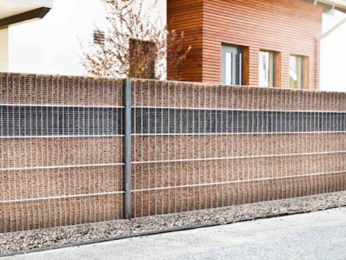 En stilfuld Gabionhegn - Panelhegn "SLEEKNOTE" 2D OVERFL.Behandl? med brune mursten og hvide metalgitterpaneler står udendørs, grus i bunden. I baggrunden ses et moderne hus med træpaneler og en hoveddør. Væggen adskiller huset elegant fra den tilstødende gade eller gangsti.