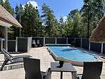 Et fredfyldt udendørs gårdhaveområde med en swimmingpool dækket med et blåt pooldæksel. Rundt om poolen er der liggestole, et lille bord med en potteplante og kurvemøbler. Området er omsluttet af et mørkt *Hegnsfabrikken* hegn, med frodige grønne træer i baggrunden under en klar blå himmel.