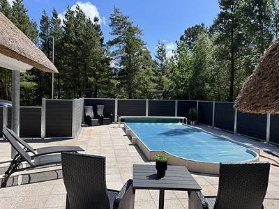 Et fredfyldt udendørs gårdhaveområde med en swimmingpool dækket med et blåt pooldæksel. Rundt om poolen er der liggestole, et lille bord med en potteplante og kurvemøbler. Området er omsluttet af et mørkt *Hegnsfabrikken* hegn, med frodige grønne træer i baggrunden under en klar blå himmel.