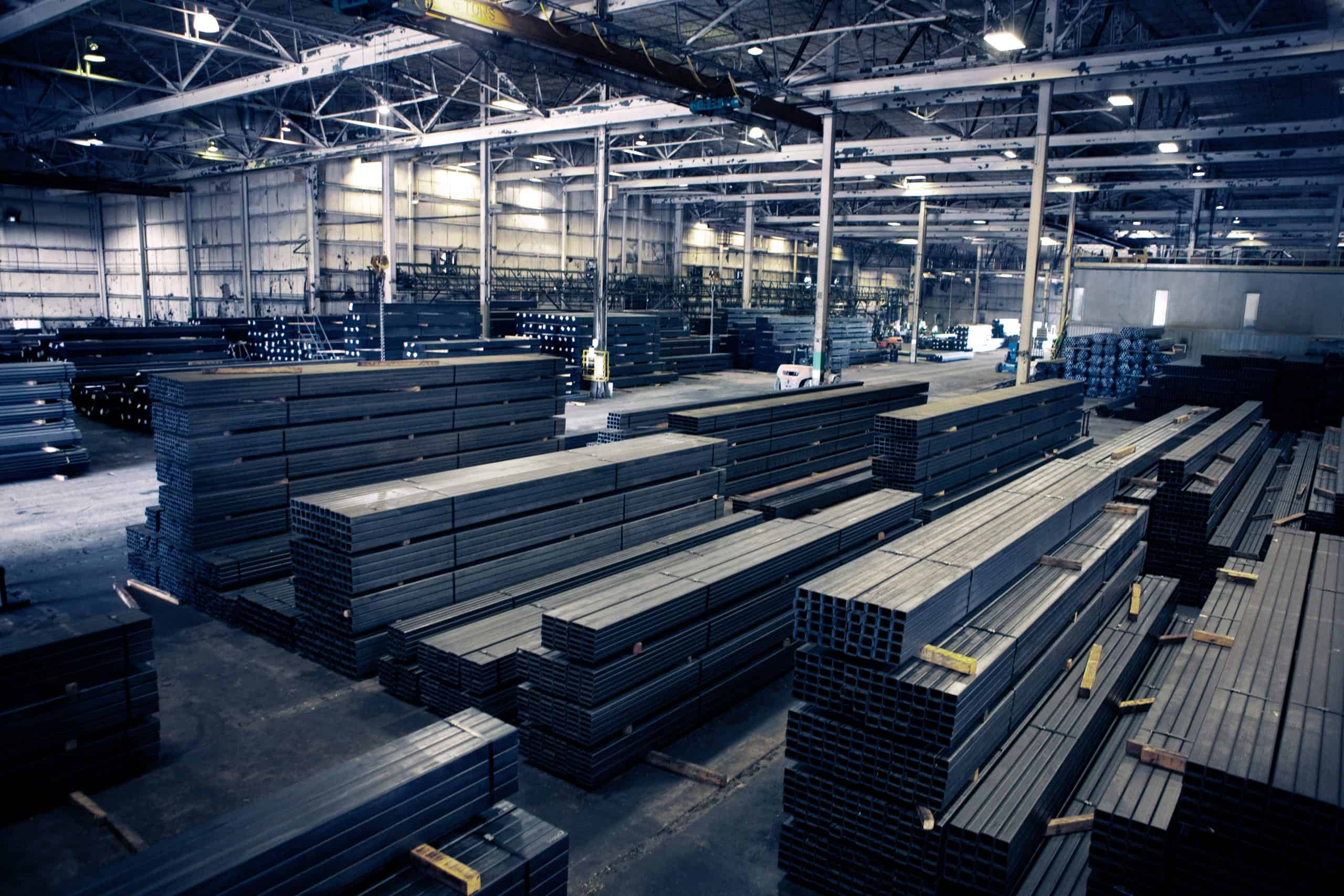 Et stort lagerinteriør fyldt med organiserede stakke af stålbjælker og metalplader. Loftet har synlige bjælker og industriel belysning. Rummet er lyst oplyst, med høje vinduer, der tillader naturligt lys at komme ind. Forskellige maskiner til overfladebehandling ses i baggrunden.