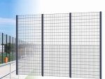 Billedet viser et højt, sort metaltrådhegn med flere lodrette og vandrette stænger, hvilket skaber et gitterlignende udseende. Dette panel hegn "BALLSTOP" 2D SPORT er installeret udendørs, med blå himmel og en del af et 2D SPORT-felt synligt i baggrunden.