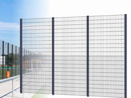 Billedet viser et højt, sort metaltrådhegn med flere lodrette og vandrette stænger, hvilket skaber et gitterlignende udseende. Dette panel hegn "BALLSTOP" 2D SPORT er installeret udendørs, med blå himmel og en del af et 2D SPORT-felt synligt i baggrunden.