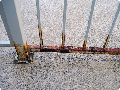 Et nærbillede af en rusten hvid metal hegn på en tekstureret betonoverflade. De nederste og lodrette stænger på hegnet er synligt korroderet med rust, og viser brun og orange misfarvning, hvilket illustrerer vigtigheden af korrekt overfladebehandling i dit materialevalg.
