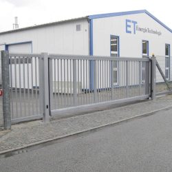 En Skydeport "STERK" skydeport med en lodret stangfacade står lukket foran en stor hvid bygning med blå trim. Bygningen, mærket "ET EnergieTechnologie," har flere store vinduer og er en del af et industrielt kompleks. Dette private anlægs sikre indgang sikrer begrænset adgang.