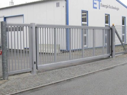 En Skydeport "STERK" skydeport med en lodret stangfacade står lukket foran en stor hvid bygning med blå trim. Bygningen, mærket "ET EnergieTechnologie," har flere store vinduer og er en del af et industrielt kompleks. Dette private anlægs sikre indgang sikrer begrænset adgang.