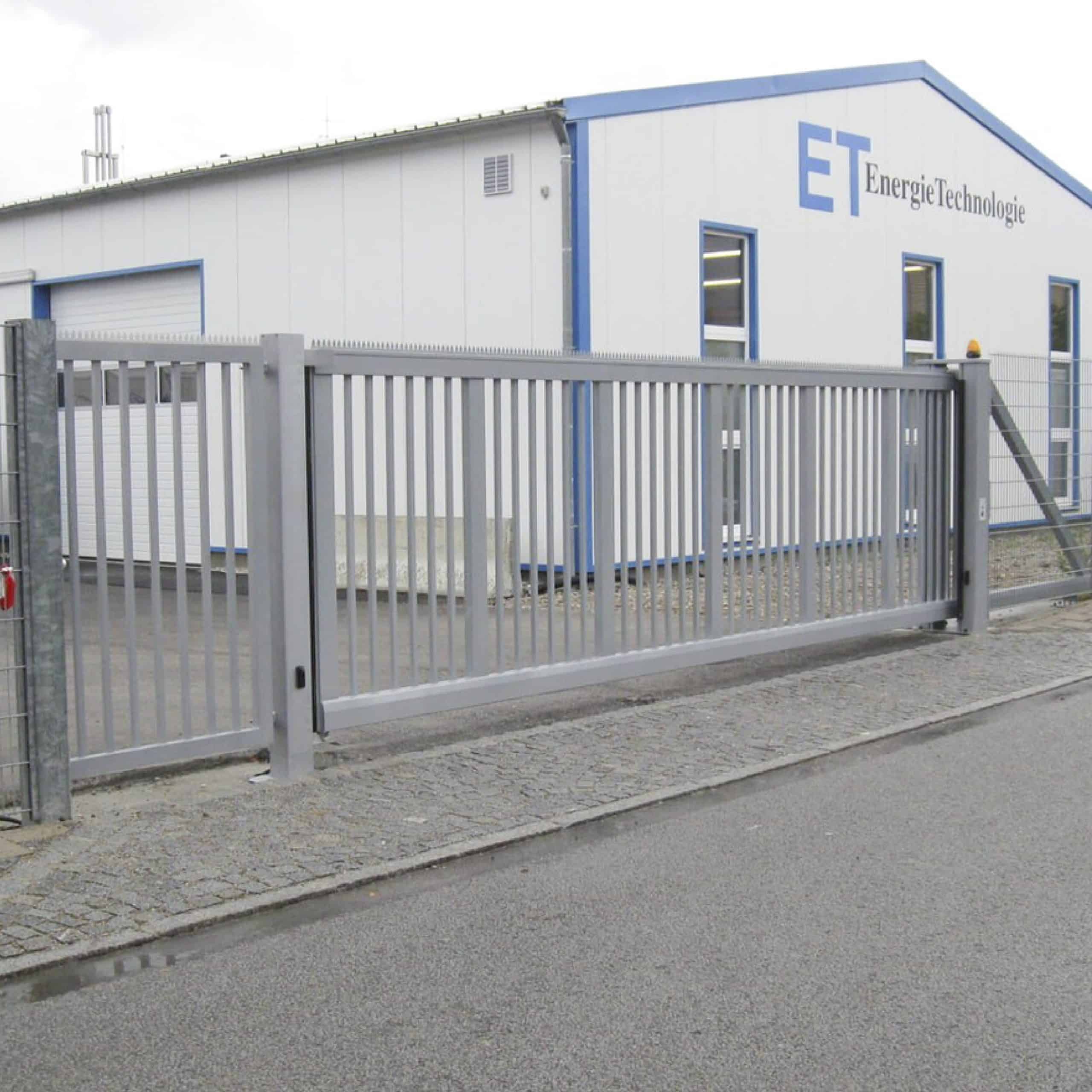 En Skydeport "STERK" skydeport med en lodret stangfacade står lukket foran en stor hvid bygning med blå trim. Bygningen, mærket "ET EnergieTechnologie," har flere store vinduer og er en del af et industrielt kompleks. Dette private anlægs sikre indgang sikrer begrænset adgang.