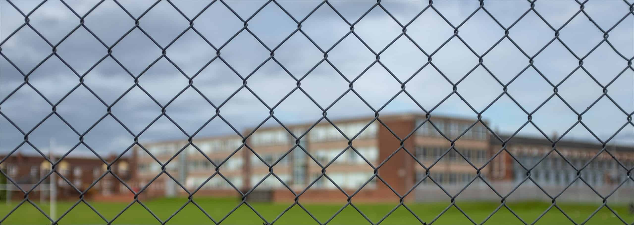 Et sløret billede af en murstensbygning med flere vinduer, set gennem et kædeled. Himlen er overskyet, og forgrunden fokuserer på det krydsende metalhegn.