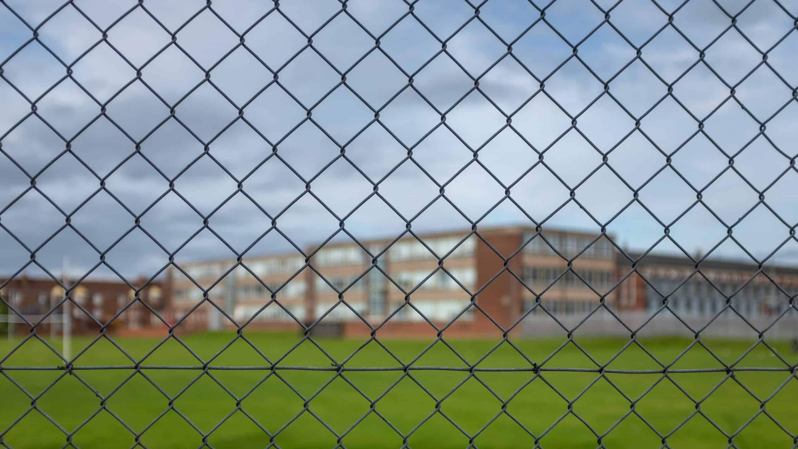 En kædehegn i forgrunden med udsigt til en stor uddannelsesinstitution i baggrunden. Bygningen er udført i røde mursten og flere vinduer. Himlen er overskyet, og der er grønt græs i grundens forhave.