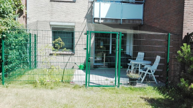 En baggård med et indhegnet terrasseområde knyttet til en murstensbygning. Terrassen har to hvide stole og et lille bord. Området, der ligner en kattevoliere, er sikret med grøn metalhegn og net strækker sig over hovedet. Nogle planter og grønne områder er synlige rundt om terrassen, hvilket fremhæver fordelene ved denne opsætning.