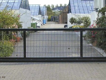 En "EasyStar"-skydeport med Lodret Gitter-design, tilgængelig i længder fra 3 til 9,5 meter og udformet til både industriel og privat brug, blokerer en asfalteret vej flankeret af grønne områder. Bag porten er drivhuse og bygninger, ledsaget af potteplanter og træer. Stien strækker sig ind i baggrunden mod en blå himmel og fjerne strukturer.