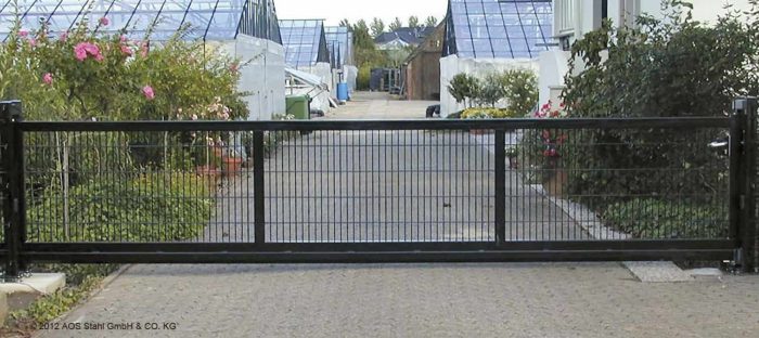 En "EasyStar"-skydeport med Lodret Gitter-design, tilgængelig i længder fra 3 til 9,5 meter og udformet til både industriel og privat brug, blokerer en asfalteret vej flankeret af grønne områder. Bag porten er drivhuse og bygninger, ledsaget af potteplanter og træer. Stien strækker sig ind i baggrunden mod en blå himmel og fjerne strukturer.